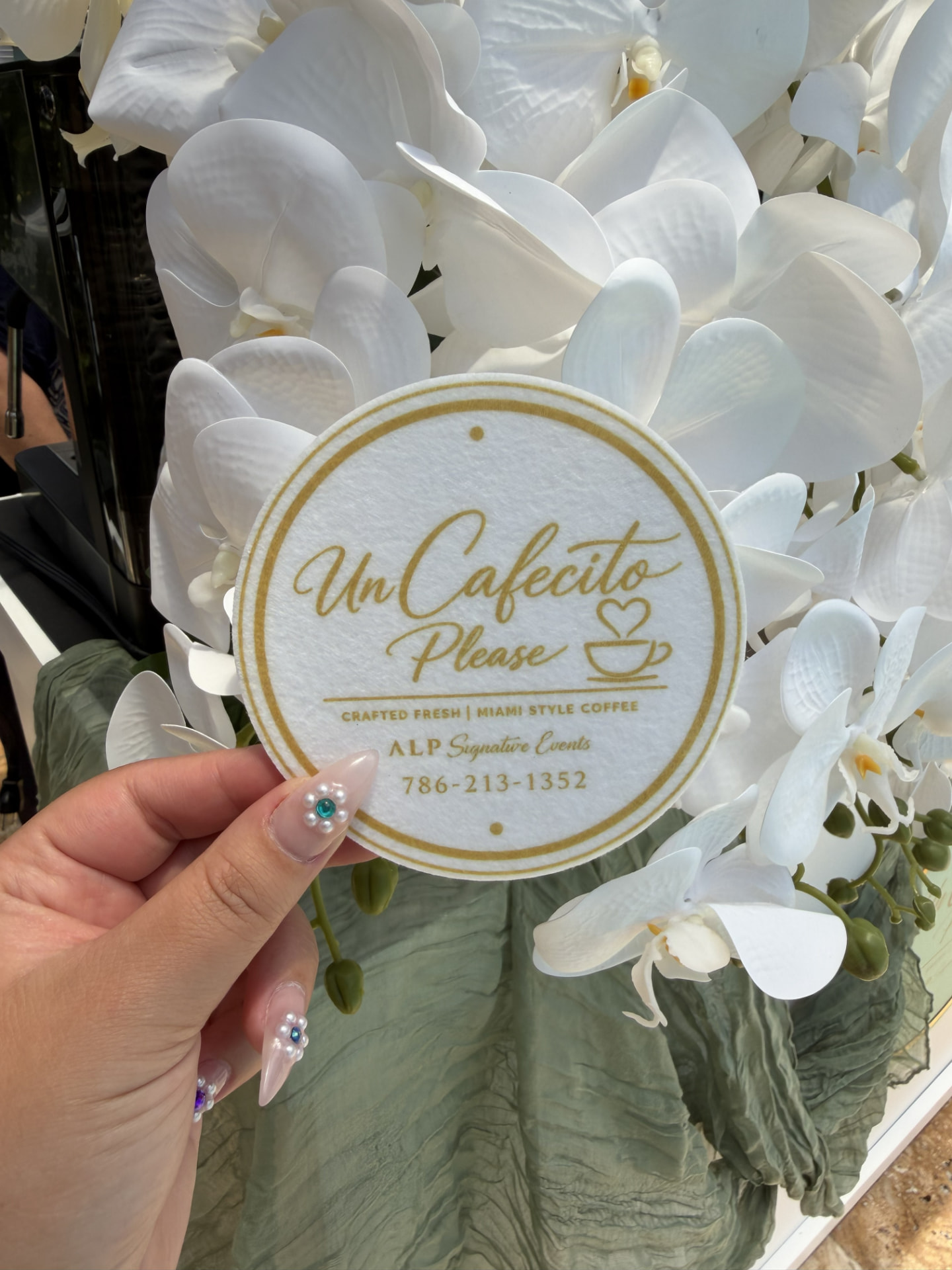Un Cafecito Please coaster held against white orchids